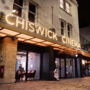 People are gathering outside the RISWICK CINEMA in the city, illuminated by the night lights and the awning of the shop across the street.
