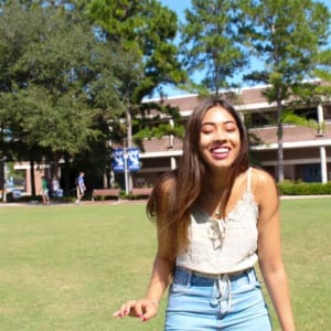 A person stands in a park, smiling and wearing a summer dress with a fashion accessory around their waist, their long hair cascading down their shoulders.