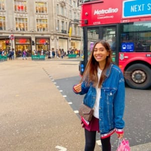 A person stands in the city wearing a red jacket, jeans, and a smile while waiting for the Metroline NEXT COMFORTDELGRO LT115 global HAM 99 LEVI'S 'S T'S bus to pass by.