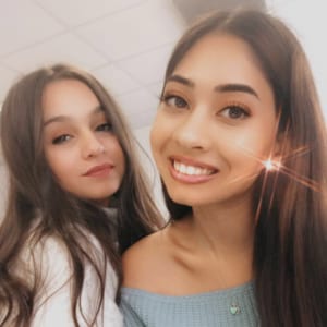 A person with long layered hair, wearing a lip gloss and a lipstick, is happily posing for a selfie with their friend, showcasing their bright smile, thick eyelashes, and beautiful irises.